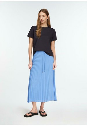 Kobieta stojąca z długimi brązowymi włosami, ubraną w czarną koszulkę, jasnoniebieską plisowaną spódnicę midi i czarne klapki na białym tle.