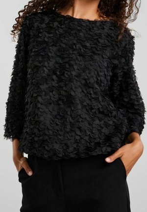 Schwarze Langarmbluse mit 3D-Blütenblattdetails, getragen zu schwarzen Hosen, Hände des Models in den Taschen, lockiges Haar an den Schultern sichtbar.