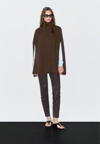 Bruine geribbelde poncho met een hoge kraag, gedragen over een lichtblauw longsleeve shirt. Gecombineerd met donkere suède broek en sandalen.