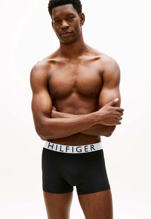 Gespiervde man staat met gekruiste armen, draagt zwarte Tommy Hilfiger boxershorts tegen een effen witte achtergrond.