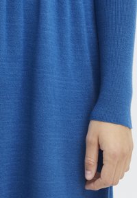 Stickad klänning i livlig blå färg, med en strukturerad tyg. Närbild visar en jogger-mudd och en hand som vilar på tyget.