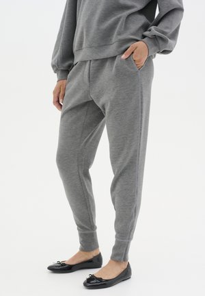Person trägt graue Jogginghose und passenden Sweatshirt mit einer Hand in der Tasche, kombiniert mit schwarzen Ballerinas vor weißem Hintergrund.