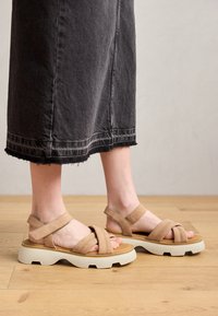Beige cross-strap sandals with a chunky white sole. Suede upper with visible stitching details. Worn with a black denim skirt.