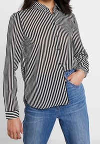 Skjorte med krave og knapper i sort og hvide lodrette striber, lange ærmer og slank pasform; kombineret med blå jeans.