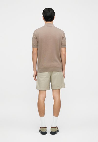 Καφέ μπλούζα πόλο με κοντά μανίκια και λεπτή υφή, σε συνδυασμό με ανοιχτό μπεζ σορτς. Το μοντέλο φορά λευκές κάλτσες και καφέ παπούτσια.