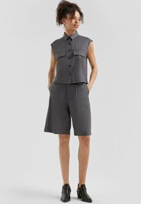 Grijze mouwloze knoopshirt met twee voorzakken, gecombineerd met knielange shorts. Gladde stof met een ontspannen pasvorm. Zwarte haklaarzen.