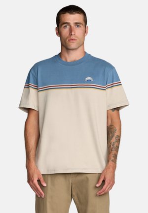 Man met kort haar, gekleed in een blauw-beige gestreept T-shirt en kaki broek, staand met zichtbare tatoeages op zijn rechteronderarm.