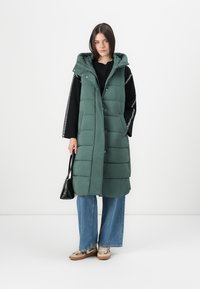 Lang grønn polstret vest med hette, med trykknapper, horisontal kviltet tekstur og sidelommer, sammen med vide jeans og en svart veske.