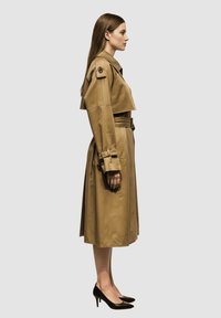 Mujer de perfil vistiendo un abrigo trinchera beige con cinturón y tacones negros sobre un fondo claro y liso.