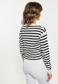 Pulover cu dungi alb-negru, dintr-un material moale, cu guler rotund, manșete și talie ribbrate. Asociat cu pantaloni albi, care evidențiază textura fundalului.