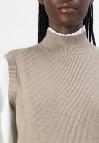 Pull à col roulé en tricot côtelé beige, au design court, avec un col montant et superposé avec une chemise blanche à volants en dessous.