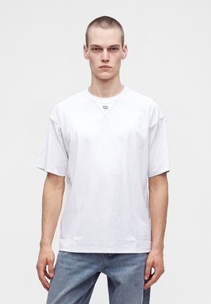 Ung mand med kort hår iført en ensfarvet, hvid oversized t-shirt og lyseblå jeans, stående mod en hvid baggrund.