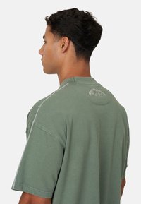 T-shirt in cotone verde con maniche corte, caratterizzato da un colletto rotondo e dettaglio ricamato sul retro. Le cuciture laterali presentano una cucitura a contrasto.