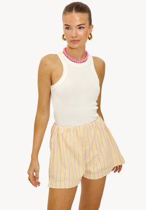 Femme portant un haut blanc sans manches à côtes, un short jaune à rayures verticales roses, un collier de perles roses, des boucles d'oreilles en anneaux dorés et une bague.