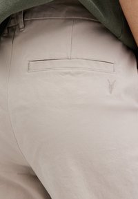 Vaaleanbeiget housut, joissa on sileä pinta, takatasku ja pieni brodeerattu emblem takataskun yläpuolella.