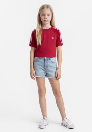 Jeune fille debout portant un t-shirt rouge à manches courtes avec des rayures blanches, un short en jean clair et des baskets blanches sur fond blanc.