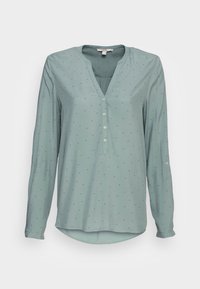 Chemise à manches longues en bleu sarcelle clair avec un col en V, une patte de boutonnage et de petits pois noirs. Tissu doux avec un ourlet arrondi.
