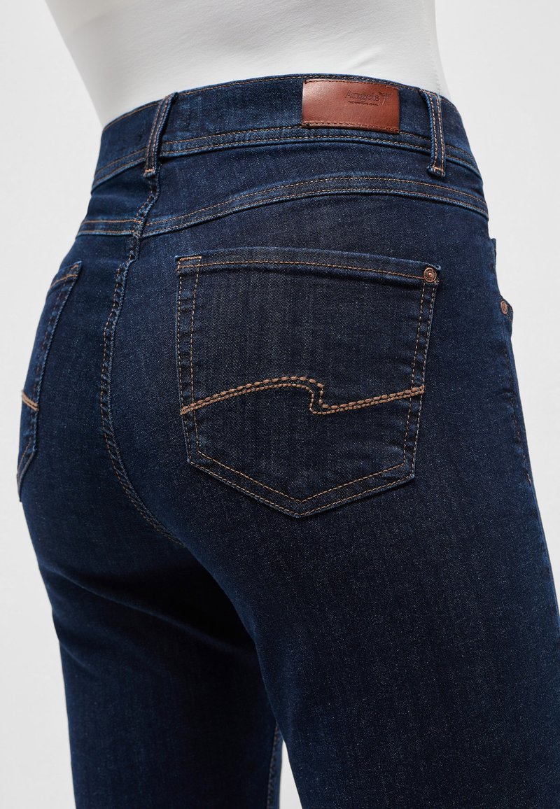 Gros plan sur un jean denim bleu foncé vu de dos, mettant en avant les poches arrière avec des coutures contrastantes et une étiquette de marque en cuir brun sur la ceinture.