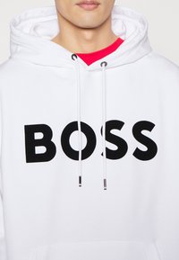 Weißer Kapuzenpullover mit großem schwarzem "BOSS"-Logo, mit Kordeln und silberner Hardware, über einem roten Hemd getragen. Glatte Stofftextur.