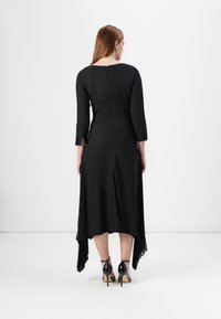 A woman in a black, long-sleeved dress with a pleated hem stands with her back to the camera, showcasing high-heeled shoes.