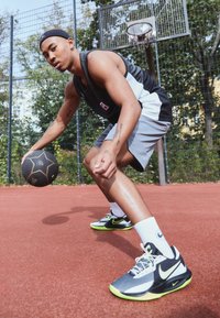 Completo sportivo da uomo con una maglietta senza maniche nera e grigia e pantaloni corti grigi. Dribbla un pallone da basket nero su un campo rosso, indossando scarpe Nike nere e verdi.