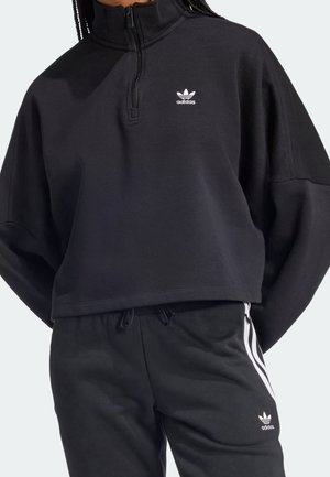 Personne portant un survêtement noir Adidas avec un sweat zippé au quart et un pantalon assorti avec des bandes blanches et le logo Adidas.