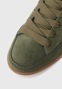 Close-up of green suede sneaker toe with wide textured laces and visible stitching on light background.