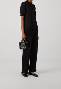 Blusa negra de manga abullonada con escote rizado, combinada con pantalones negros con puntos y un pequeño bolso negro con un acento de flor texturizada.