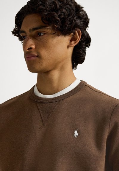 Brązowa bawełniana bluza z okrągłym dekoltem i białym logo. Charakteryzuje się miękką fakturą i raglanowymi rękawami, noszona na białej koszuli.