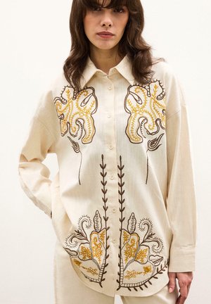 Femme portant une chemise crème à boutons avec manches longues, ornée d'une broderie florale jaune et brune sur le devant et les manches.