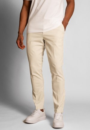Hombre que lleva pantalones ajustados beige, zapatillas blancas y una camiseta beige claro, de pie con una mano en el bolsillo frente a un fondo liso.