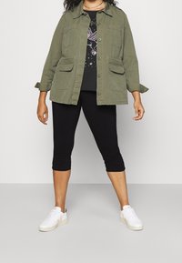 Viheroliivi takki, jossa on kaksi etupussia, päällä musta graafinen t-paita, mustat lyhyet legginsit ja valkoiset tennarit vaaleanpunaisilla yksityiskohdilla.