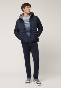 Marineblaue Daunenjacke mit Kapuze, grauem Innenfutter, blau gestreiftem Pullover darunter, dunklen Hosen und weißen Sneakern mit blauen Akzenten.