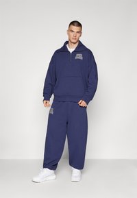 Sudadera azul marina con cuello de media cremallera y bolsillo frontal, combinada con pantalones de chándal holgados a juego. Zapatillas blancas completan el atuendo.