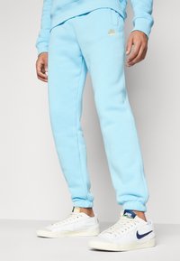 Ljusblå sweatpants med en figursydd design, elastiska muddar, sidofickor och en liten gyllene Nike-logotyp på vänster lår.