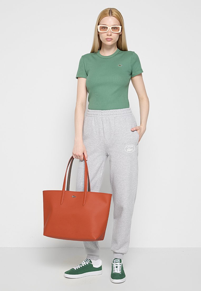 Zielona, prążkowana koszulka, szare dresowe spodnie z logo, czerwona skórzana torba oraz zielone sneakersy. Jednolity tle, model stoi z rękami w kieszeniach.