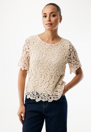 Beige kanten top met korte mouwen en gegolfde randen, met een bloemenpatroon en opengewerkt materiaal, gedragen met donkere spijkerbroek.