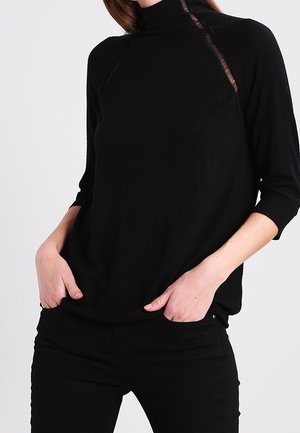 Osoba ubrana w czarny golf z rękawami trzy czwarte i czarne spodnie, ręce w kieszeniach, widoczna od ramion do bioder.