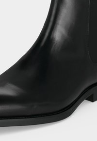 Bottine noire en cuir avec finition lisse, bout arrondi, pan élastique sur le côté et petit talon épais sur fond blanc.