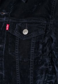 Tmavě modrá sametová bunda se strukturálním designem, která má náprsní kapsu, stříbrný patent a červenou štítkovou značku Levi's. Texturovaná látka.