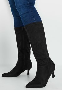 Botas de ante negras hasta la rodilla con punta afilada y un tacón pequeño, que cuentan con una cremallera lateral para el cierre y una textura suave.