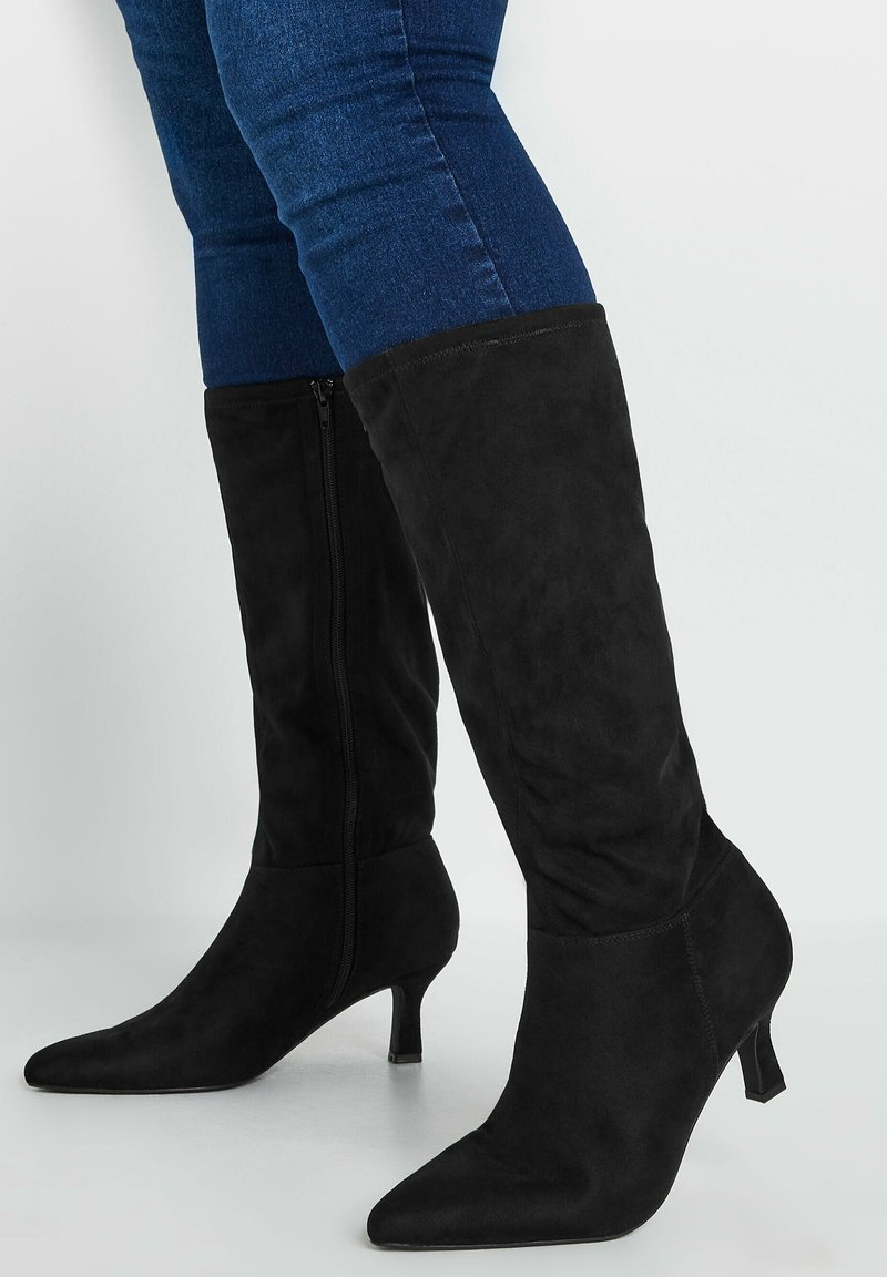 Botas de ante negras hasta la rodilla con punta afilada y un tacón pequeño, que cuentan con una cremallera lateral para el cierre y una textura suave.
