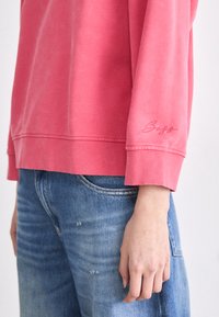 Sudadera rosa de manga larga con puños acanalados. Tiene un ajuste casual y un logo bordado en la manga. La lleva puesta con jeans azules desgastados.