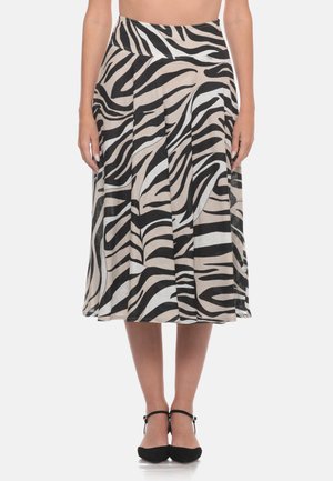 Donna che indossa una gonna a vita alta, lunga fino al ginocchio, con righe zebra nere e beige e scarpe nere con cinturino alla caviglia, in piedi davanti a uno sfondo semplice.