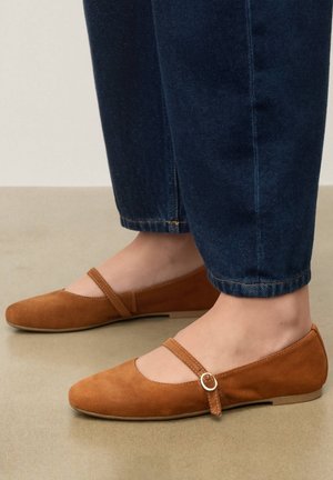 Személy barna szarvasbőr Mary Jane balerinacipőben, csatos pántokkal és sötétkék farmernadrágban, bézs padlón állva.