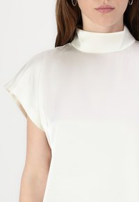 Haut blanc sans col avec une texture lisse et brillante et des manches courtes retroussées, présentant un design simple et épuré sans motifs ni accents.