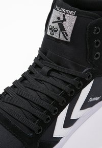 Black high-top sneakers featuring textured fabric, laces, a white logo, and contrasting white zigzag accents on the side.