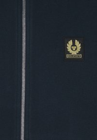 Marinblå zip-up sweatshirt gjord av mjukt tyg, med en silverfärgad dragkedja och en svart lappar med guldigt logo på vänster bröst.