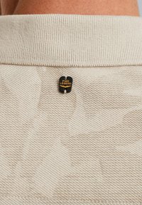 Polo en tissu beige texturé avec un col à côtes, arborant une petite étiquette noire et dorée PME Legend à l'arrière près de l'encolure.
