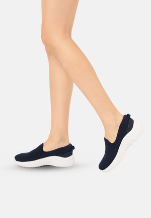 Persoon draagt marineblauwe slip-on sneakers met witte zolen en loopt op een effen lichte achtergrond.